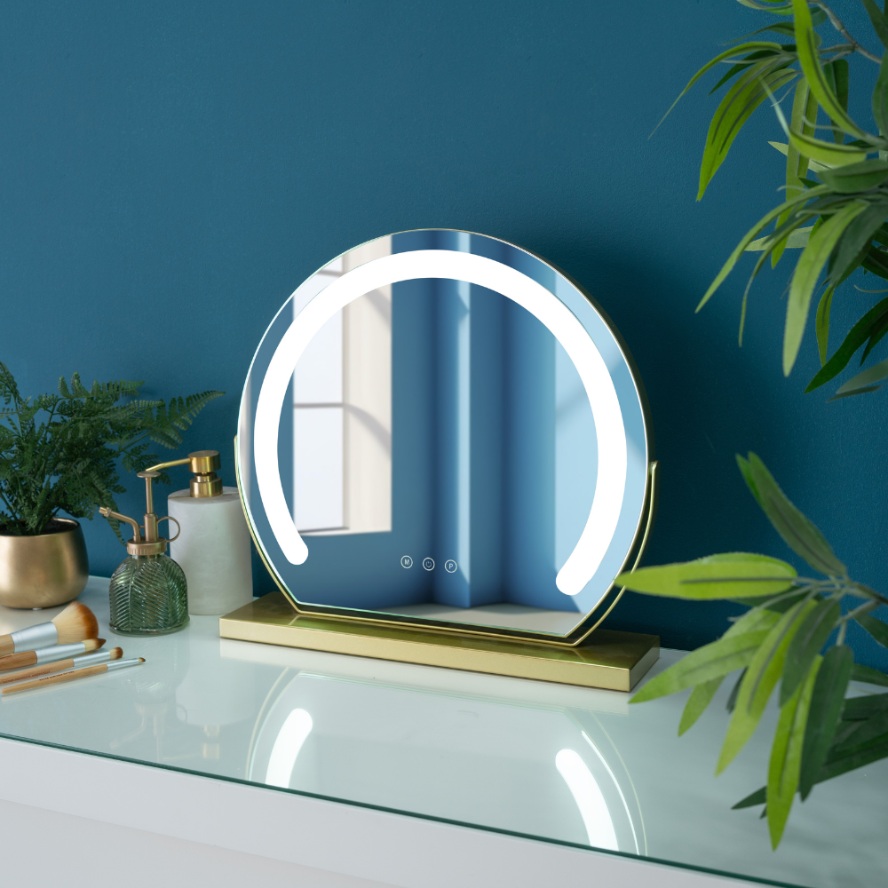 A circular light up LED makeup mirror with a bronze base, placed on a dressing table and surrounded by bottles and beauty products